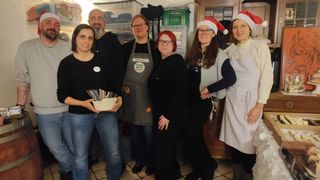 Eine Gruppe von sechs Personen, die Schürzen und Weihnachtsmützen tragen, posiert für ein Foto. Einer hält eine Schüssel mit Gegenständen. Sie stehen in der Nähe von Lagerboxen und einem Holzschrank.