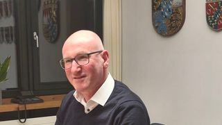 A man wearing glasses sits in an office chair, smiling. Behind him is a desk with a telephone. On the wall are two shields with emblems. A plant is on the left.