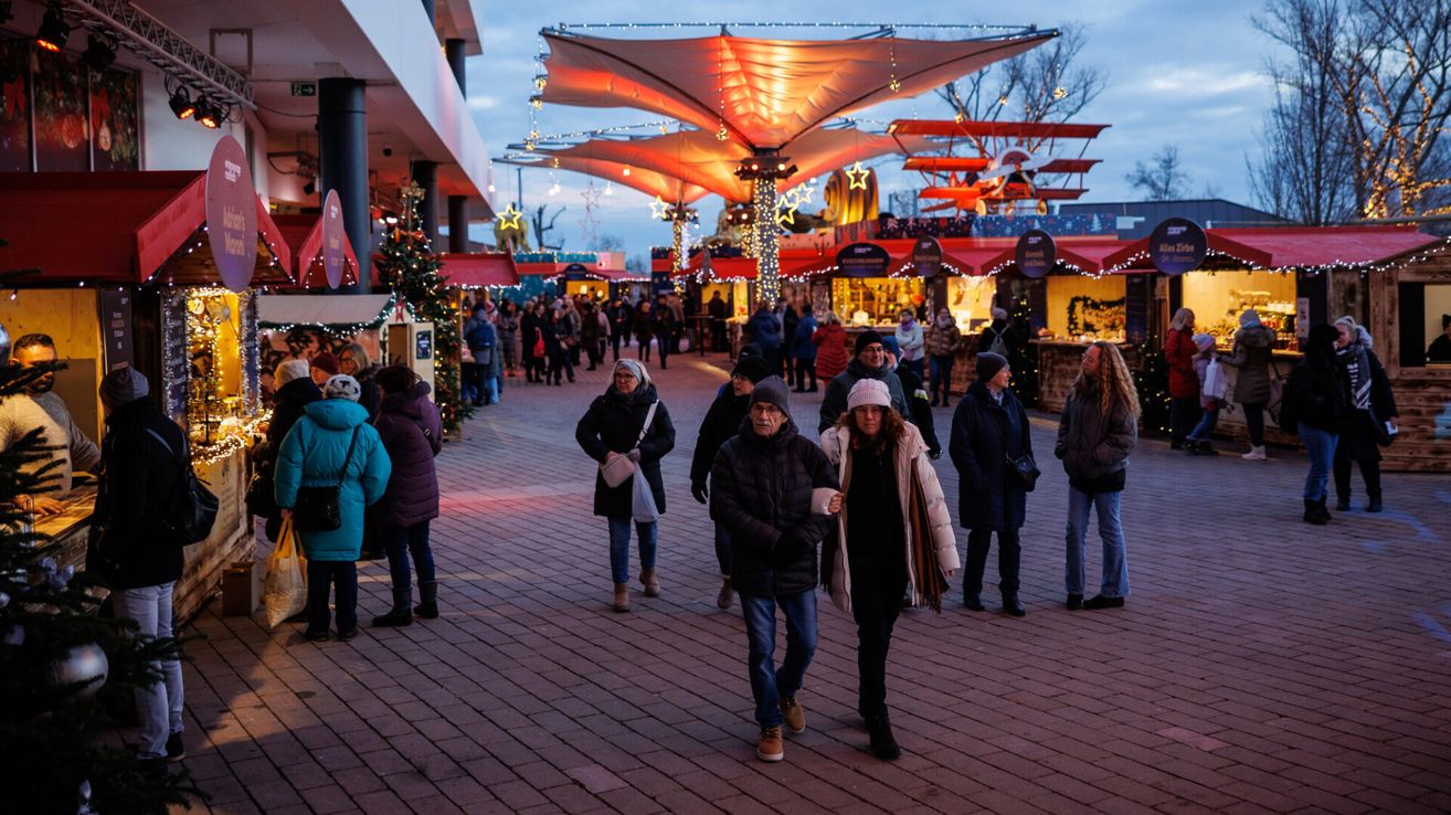 Ein geschäftiger Freiluftmarkt mit festlicher Atmosphäre, Menschen gehen durch die Stände. Es ist Nacht, und der Bereich ist mit Weihnachtsbeleuchtung und Bäumen geschmückt.