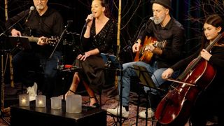 Eine kleine Band spielt mit einer singenden Frau und einem Mann, der Gitarre spielt, vor einem Mikrofon sitzend. Ein anderer Mann spielt Cello und ein weiterer Gitarre.