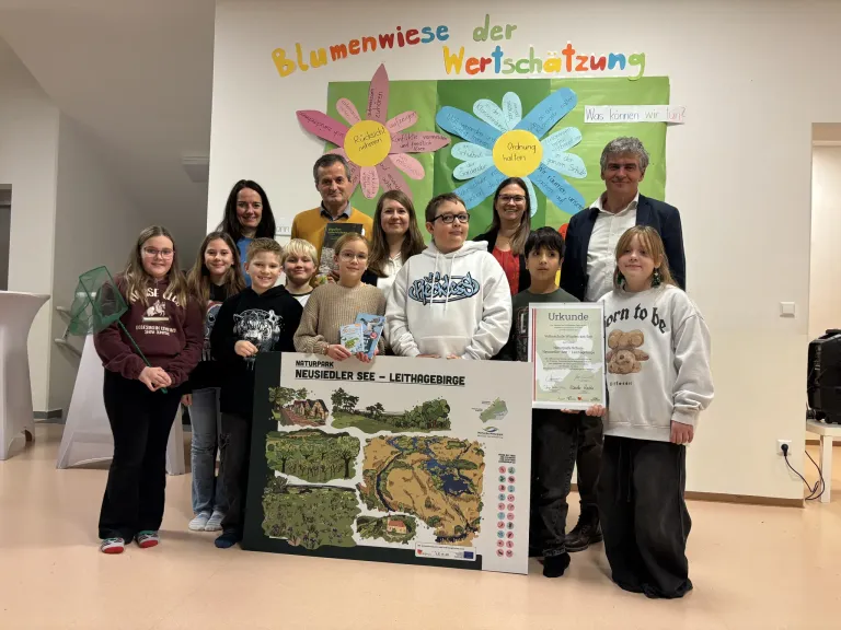 Gruppenfoto von Erwachsenen und Kindern vor einem Hintergrund mit dem Titel 'Blumenweise der Wertschätzung', wobei zwei Kinder ein Zertifikat und eine Karte halten.