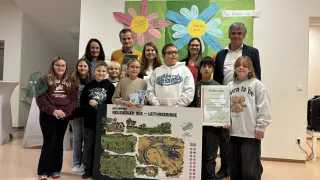 Gruppenfoto von Erwachsenen und Kindern vor einem Hintergrund mit dem Titel 'Blumenweise der Wertschätzung', wobei zwei Kinder ein Zertifikat und eine Karte halten.