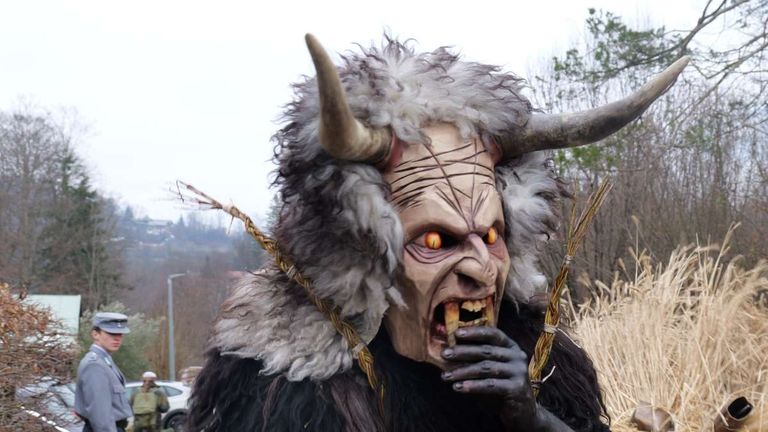 A person dressed as a scary horned demon with an open mouth and a tree branch in their hand is standing in a field.