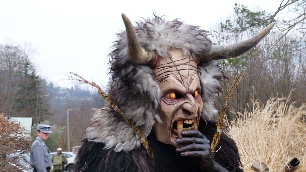 A person dressed as a scary horned demon with an open mouth and a tree branch in their hand is standing in a field.