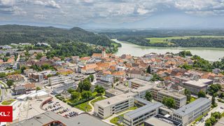 Aerial view of a town with buildings, a river, and green hills. The town has many buildings with orange roofs, and a prominent church with a tower. The river runs through the town and is surrounded by greenery. The sky is cloudy.
