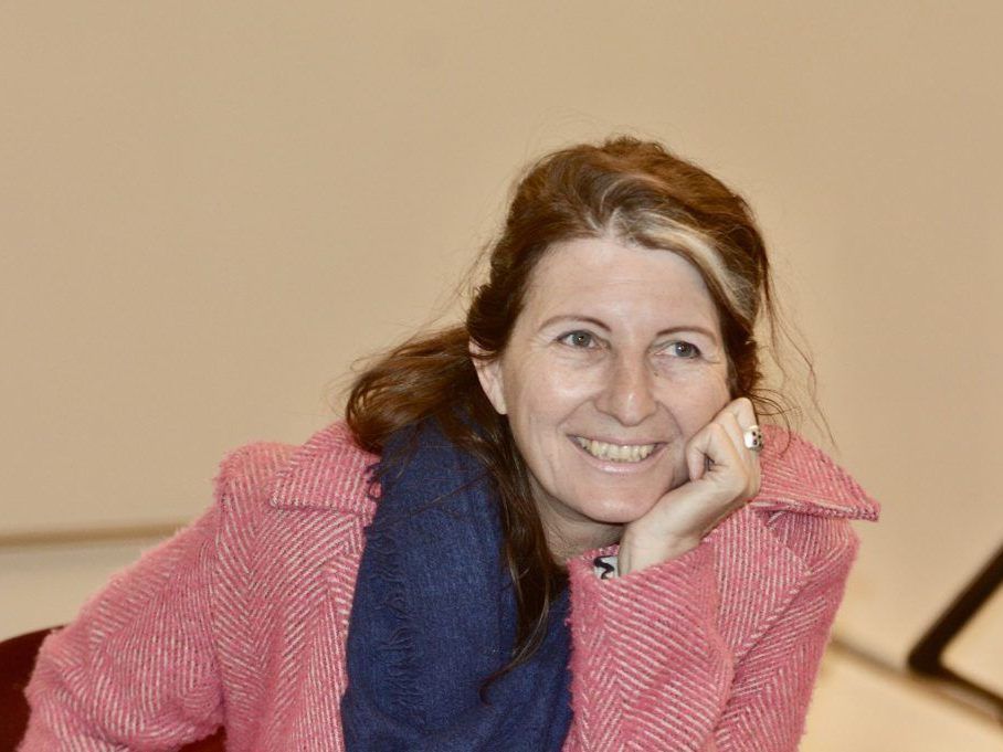 Eine Frau mit braunen Haaren und einem Lächeln im Gesicht sitzt und posiert für ein Foto. Sie trägt einen rosa Mantel und einen blauen Schal.