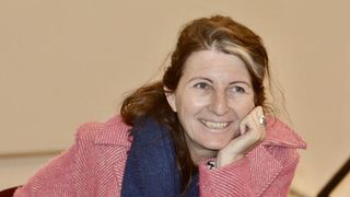 Eine Frau mit braunen Haaren und einem Lächeln im Gesicht sitzt und posiert für ein Foto. Sie trägt einen rosa Mantel und einen blauen Schal.