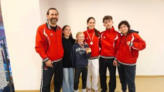 Eine Gruppe junger Personen und ein Mann posieren für ein Foto. Sie tragen alle rote Jacken und haben Medaillen um den Hals. Der Mann trägt eine rote Jacke mit einem Logo auf der linken Seite.