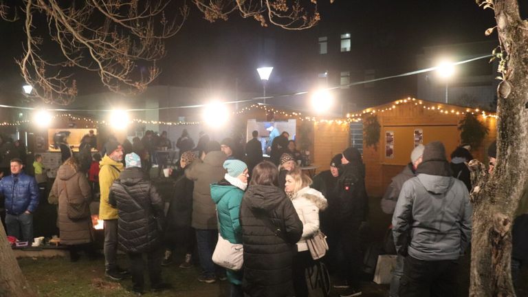 Eine Gruppe von Menschen steht draußen im Dunkeln, in der Nähe von Holzhütten mit Lichterketten. Sie sind warm gekleidet, wahrscheinlich in einer Warteschlange für eine Veranstaltung. Der Bereich ist von Lampen beleuchtet.