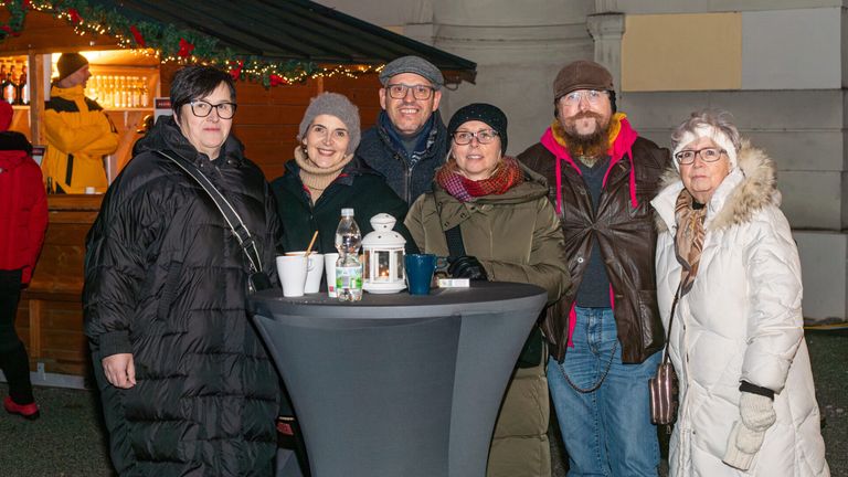 Fünf Personen posieren für ein Foto neben einem Tisch mit Tassen und einer Laterne. Sie tragen Winterkleidung. Hinter ihnen befindet sich ein Holzstand mit Weihnachtsdekoration.