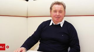 Ein Mann mit grauem Haar trägt einen marineblauen Pullover mit weißem Kragen und sitzt in einer Sitznische mit einer weißen Wand dahinter.