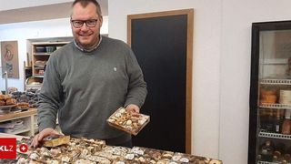 Ein Mann mit Brille, der eine Box mit Nüssen und getrockneten Früchten hält, steht vor einer weißen Wand mit einer Tafel. Er lächelt und scheint sich am Arbeitsplatz zu befinden.