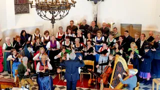 Ein Chor singt in einer Kirche mit einem Dirigenten vorne. Viele Musiker spielen Instrumente. Ein Kronleuchter hängt von der Decke.