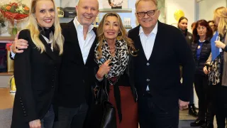 Vier Menschen in formeller Kleidung stehen eng zusammen für ein Foto und lächeln. Eine Frau in der Mitte trägt einen Leopardenprint-Schal. Ein Mann rechts trägt eine Brille. Eine Frau links hat lange blonde Haare. Im Hintergrund halten Regale verschiedene Gegenstände.