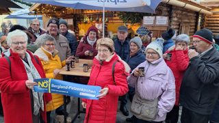 A group of people in winter clothes are standing in front of a food stall. They are holding cups and seem to be posing for a photo. The stall has a wooden table with cups on top. There are hanging items on the ceiling. The area is surrounded by wooden walls with Christmas decorations.