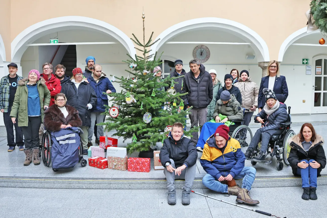 Eine Gruppe von Menschen versammelt sich um einen geschmückten Weihnachtsbaum mit Geschenken, einige sitzen und andere stehen, einige in Rollstühlen. Sie tragen Winterkleidung und Mützen.