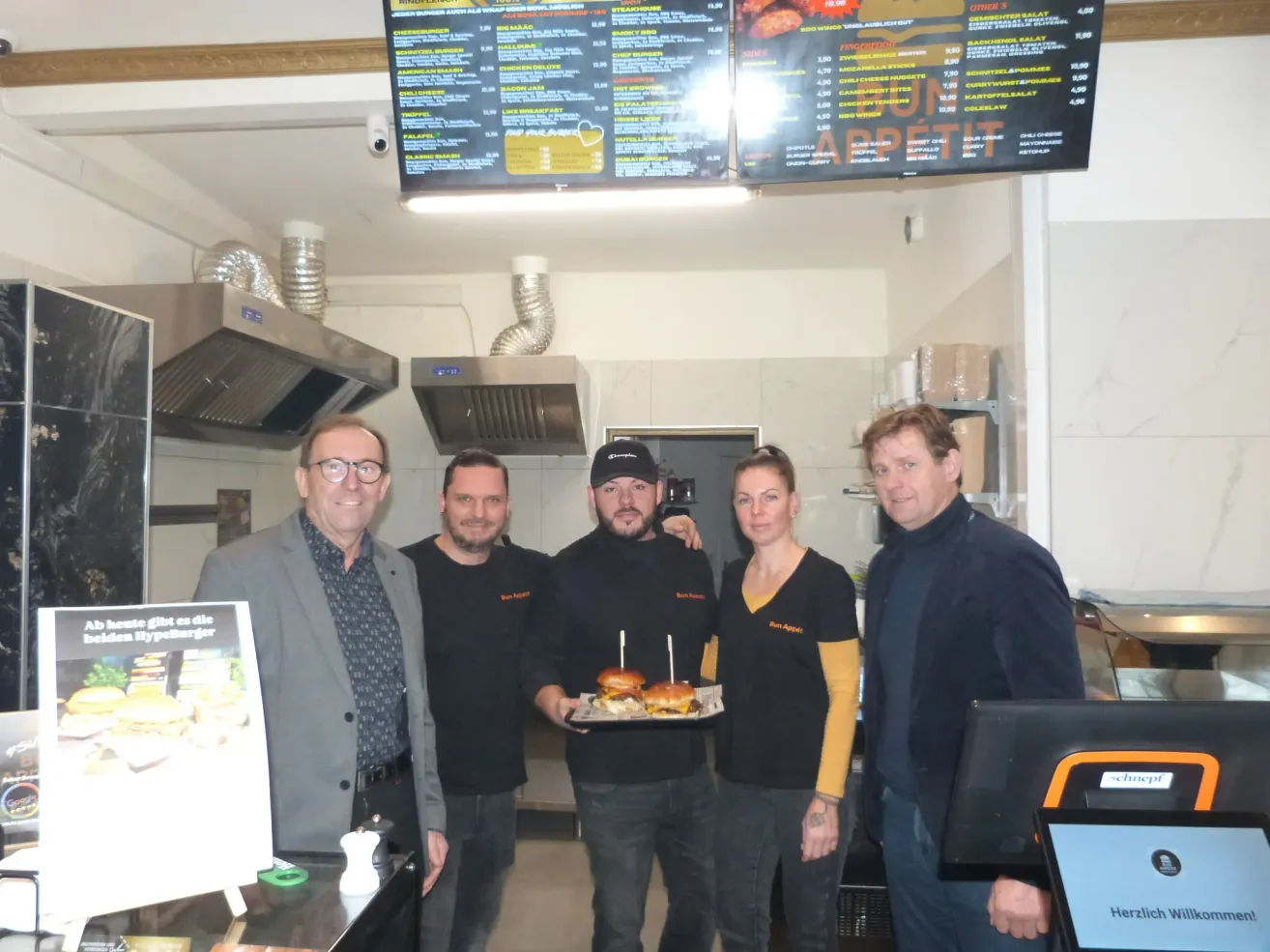 In einem Restaurant steht eine Gruppe von Menschen zusammen. Ein Mann hält einen Tablett mit Sandwiches, während andere lächeln und in die Kamera schauen. Ein Menübrett ist über ihnen, und Küchengeräte sind im Hintergrund sichtbar.