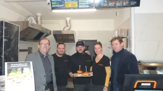 In einem Restaurant steht eine Gruppe von Menschen zusammen. Ein Mann hält einen Tablett mit Sandwiches, während andere lächeln und in die Kamera schauen. Ein Menübrett ist über ihnen, und Küchengeräte sind im Hintergrund sichtbar.