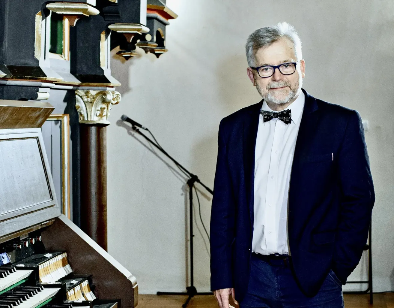 Ein älterer Mann in Anzug und Fliege steht neben einem Mikrofon vor einer Orgel.
