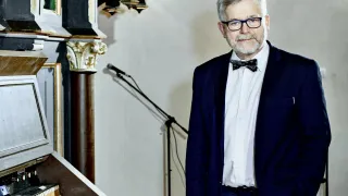 Ein älterer Mann in Anzug und Fliege steht neben einem Mikrofon vor einer Orgel.