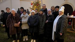Eine Gruppe von Menschen in Winterkleidung, die vor einem geschmückten Weihnachtsbaum mit brennenden Kerzen stehen.