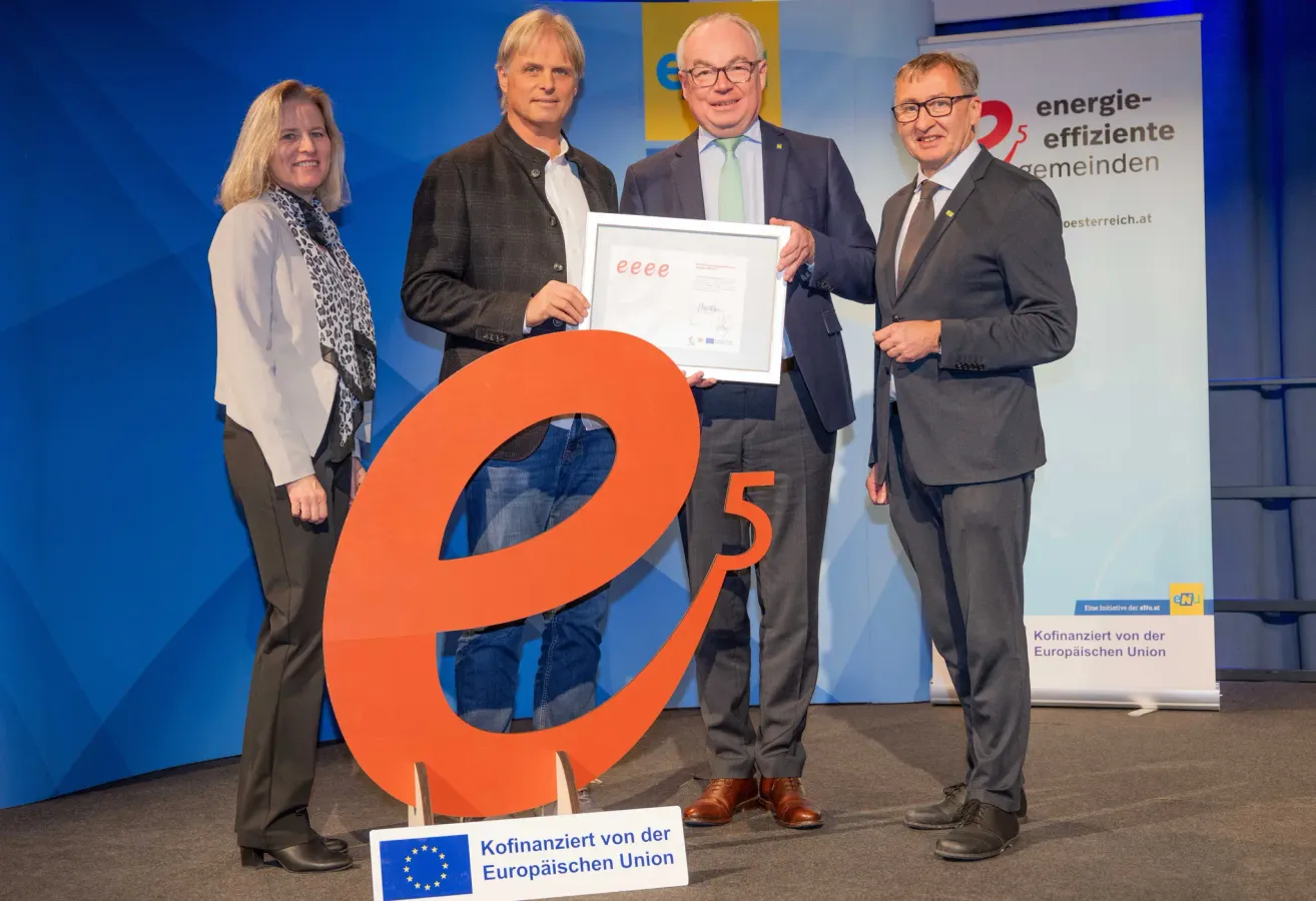 Vier Personen stehen vor einer großen orangefarbenen Nummer 5 und halten ein Zertifikat. Hinter ihnen ist eine blaue Kulisse mit einem Logo und Text.