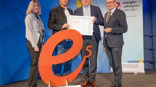 Vier Personen stehen vor einer großen orangefarbenen Nummer 5 und halten ein Zertifikat. Hinter ihnen ist eine blaue Kulisse mit einem Logo und Text.