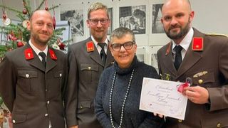 Eine Frau in einem grauen Pullover steht lächelnd und hält eine Urkunde, umgeben von drei Männern in Uniformen mit einem Weihnachtsbaum im Hintergrund.