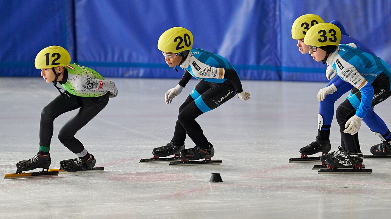 Drei Eisläufer mit Helmen und Schlittschuhen stehen in einer Reihe auf einer Eisbahn. Der Läufer in der Mitte hat die Nummer 20 auf seinem Helm.