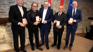 Fünf Männer in Anzügen stehen nebeneinander und halten Auszeichnungen. Sie tragen alle formelle Kleidung, zwei von ihnen tragen Brillen. Hinter ihnen sind drei Fahnen an einer Steinmauer befestigt.