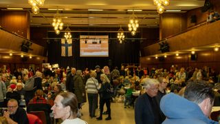 Ein großer Raum beherbergt ein geselliges Treffen mit vielen Menschen, die sich unterhalten. Eine Projektionsleinwand zeigt 'Mumin Jagd'. Eine finnische Flagge hängt auf der linken Seite.