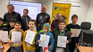 A group of boys and adults pose for a photo, holding certificates. One boy wears a beanie. Behind them, a chessboard is displayed on a screen.