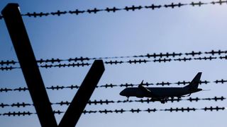 Eine Silhouette eines Flugzeugs fliegt über Stacheldrahtzäune unter einem blauen Himmel.