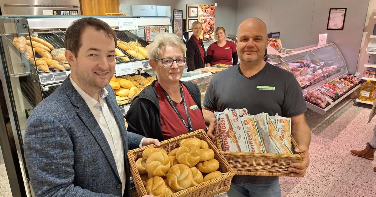 Drei Menschen lächeln und posieren für ein Foto in einer Bäckerei, mit Brotkörben und Gebäck vor ihnen. Dahinter befindet sich ein Glasvitrine, gefüllt mit verschiedenen Gebäcksorten.