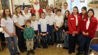 Eine Gruppe von Kindern und Erwachsenen posiert für ein Foto. Sie stehen in einem Raum mit Holzböden. Einige von ihnen tragen Brillen. Der Mann in der Mitte ist als Weihnachtsmann gekleidet. Hinter ihnen stehen Regale mit Topfpflanzen und anderen Gegenständen. In der Nähe des Glasfensters steht eine Person.