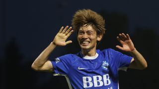 A soccer player celebrates with both hands raised in the air, wearing a blue jersey with white letters and the Nike logo.