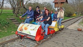 Eine Gruppe von Menschen fährt auf einem pedalbetriebenen Zug auf einer Schiene, wobei eine Frau in die Pedale tritt und andere lächeln. Der Zug ist mit Kürbissen und einem VW-Logo verziert. Hinter ihnen ist ein Haus mit rotem Dach und einer Rutsche zu sehen.