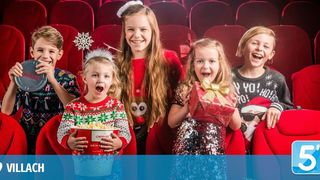 Five children are seated in a theater with red seats, smiling and holding gifts and popcorn. The boy on the left holds a silver disc. The girl on the right wears a Santa hat.