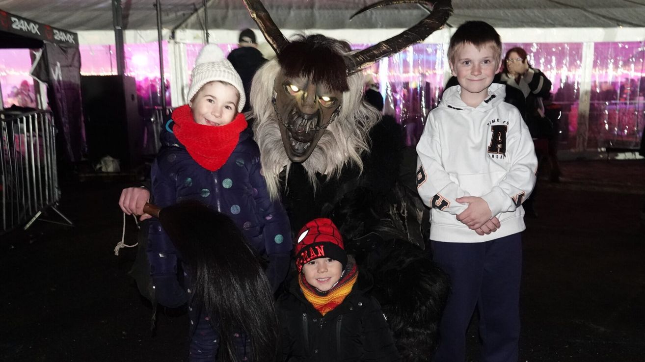 Drei Kinder, eines mit einer Spiderman-Mütze, posieren mit einer Person in einer gruseligen Kostümierung mit Hörnern und einer Maske. Sie stehen nebeneinander.