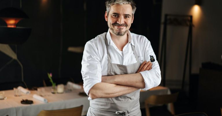 Ein Koch in einem weißen Hemd und einer grauen Schürze mit verschränkten Armen in einem Restaurant. Hinter ihm steht ein Tisch mit einer Blume und Kerzen.