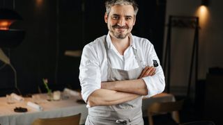 Ein Koch in einem weißen Hemd und einer grauen Schürze mit verschränkten Armen in einem Restaurant. Hinter ihm steht ein Tisch mit einer Blume und Kerzen.