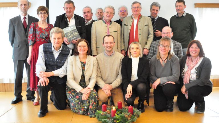 Eine Gruppe von Menschen posiert für ein Foto, einige kniend und andere stehend. Ein Mann hält ein Buch, und drei brennende Kerzen stehen im Vordergrund.