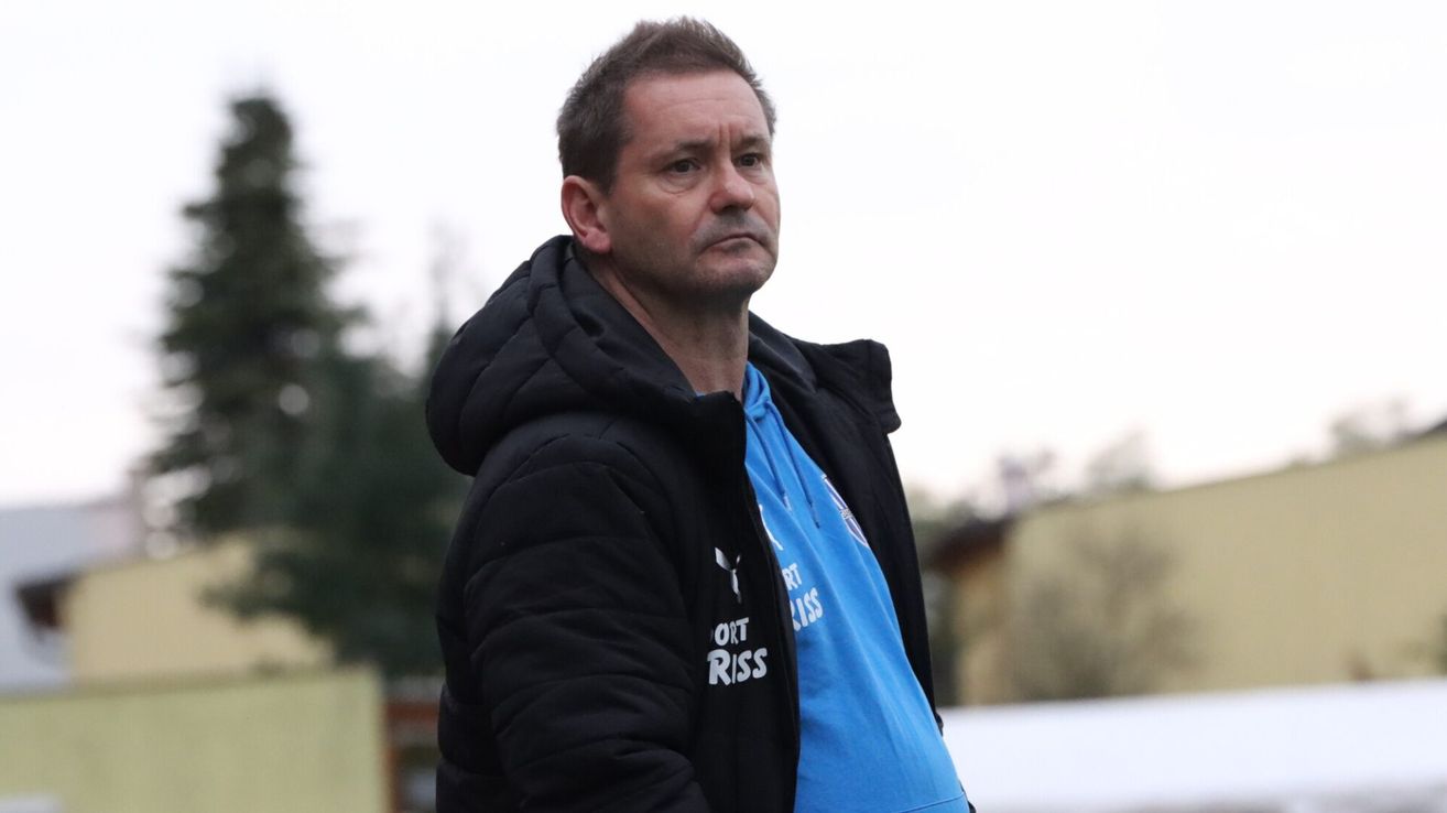 Ein Mann in einem blauen Shirt mit weißem Text auf einem schwarzen Jackett steht im Freien und schaut zur Seite.