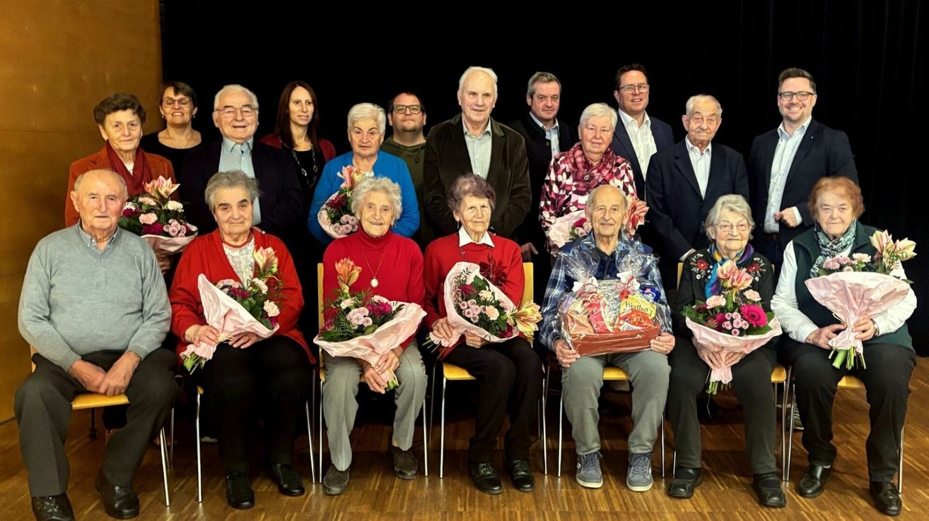 Eine Gruppe älterer Menschen posiert für ein Foto, während sie Blumen und Geschenke halten. Sie sitzen auf Stühlen vor einem Holzboden.