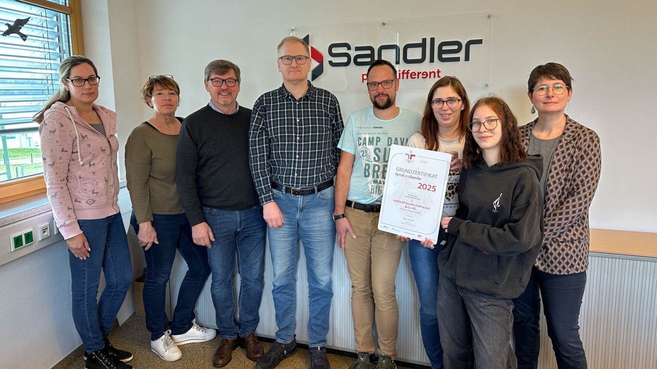A group of six individuals stands in front of a white backdrop with the word Sandler written in red. They are holding a certificate for 'Grundzertifikat berufsfamilie 2025.' They are all wearing glasses and shoes.
