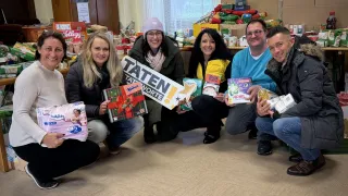 A group of people are smiling and posing for a photo. They are holding gifts and boxes, and some of them are wearing hats. The boxes have the word 'Taten' on them. There are also boxes of food on the table behind them. There are posters on the wall, and there is a window with a curtain.