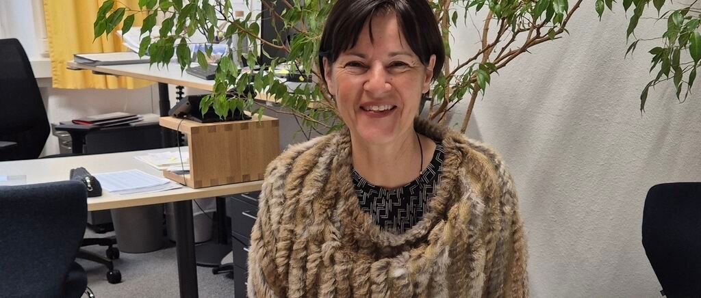 A woman with short hair and a fur scarf is smiling while standing in front of a desk with a plant.