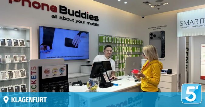 Ein Smartphone-Laden mit einem großen Bildschirm, der ein Handy zeigt, einem Mann am Tresen und einer Frau, die einen roten Gegenstand hält. Mehrere Telefone sind in Glasvitrinen ausgestellt.