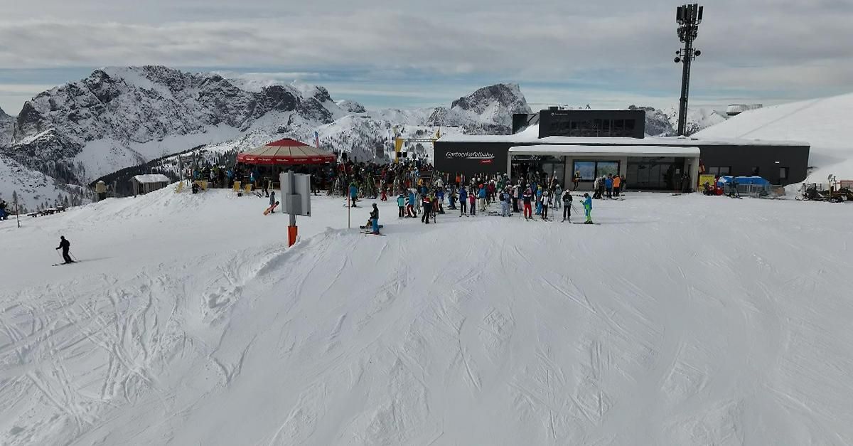 Ein überfüllter Skibereich mit vielen Skifahrern auf einem verschneiten Hang, einem roten Vordach und einem Gebäude im Hintergrund.