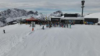 Ein überfüllter Skibereich mit vielen Skifahrern auf einem verschneiten Hang, einem roten Vordach und einem Gebäude im Hintergrund.